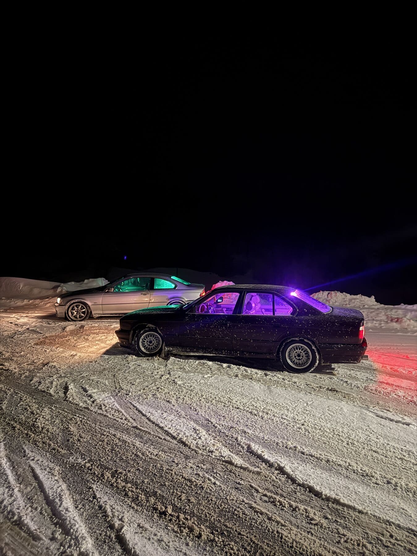 Zwei BMW nachts auf Schnee, einer mit lila Innenbeleuchtung, mit Blitz fotografiert